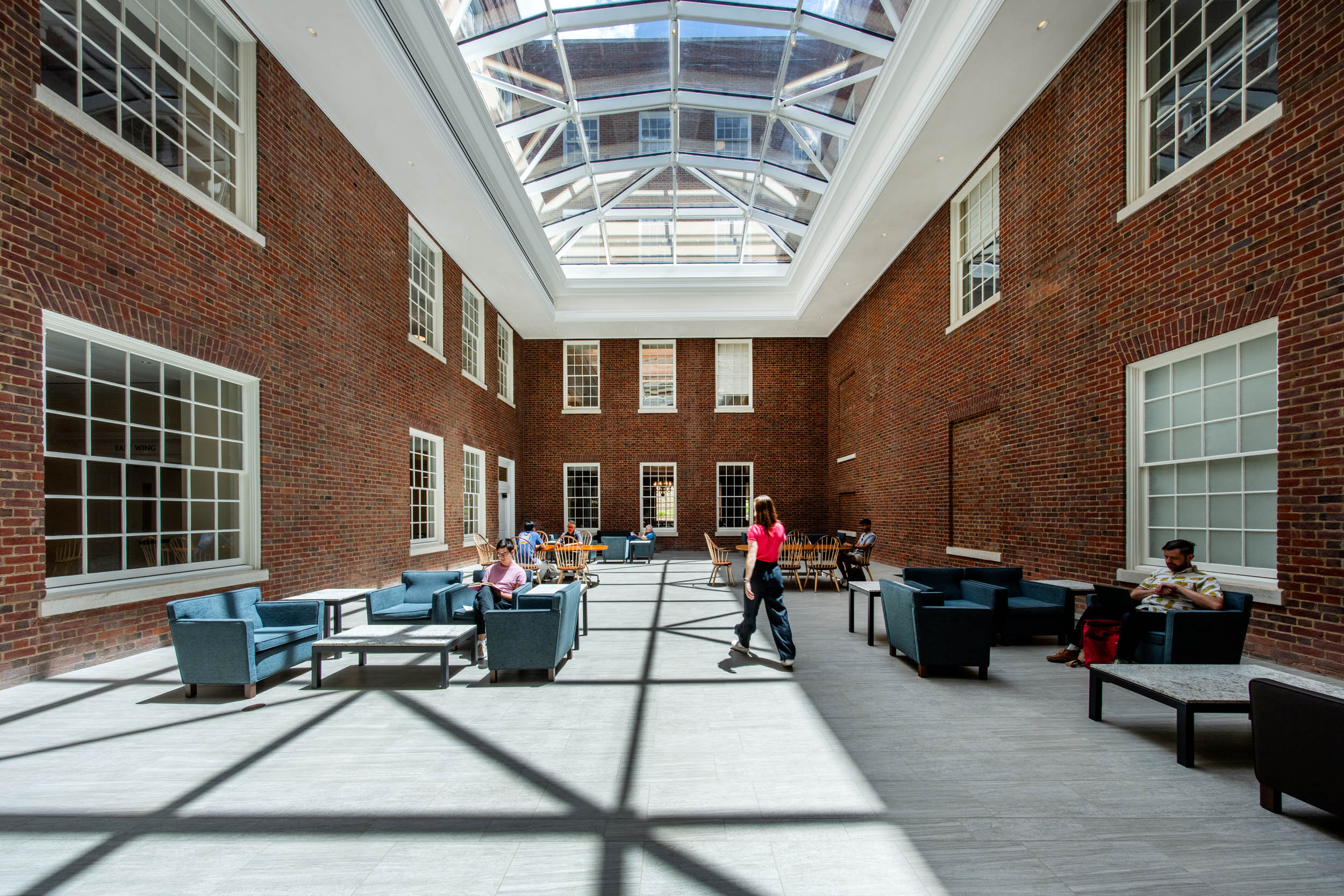 Interior of a spacious atrium with brick walls, large windows, and a glass ceiling. Several individuals are seated at tables, studying and interacting. Comfortable seating arrangements are spread throughout the space.