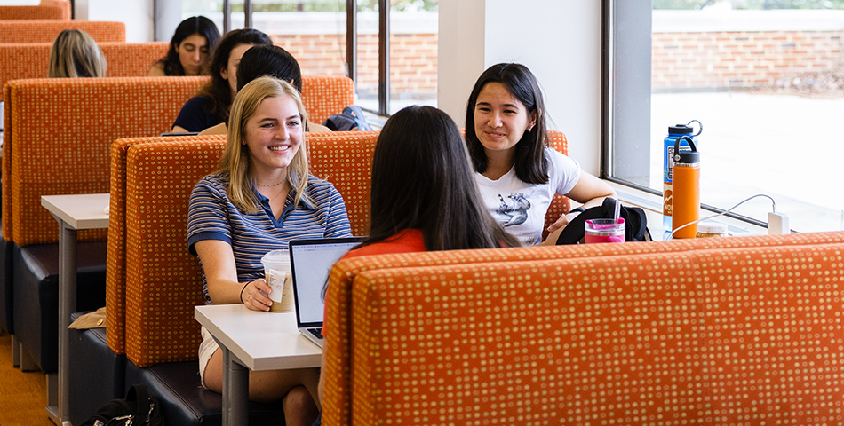 Students sit in a window-side booth, with laptops on the table. They are smiling and appear mid-conversation.
