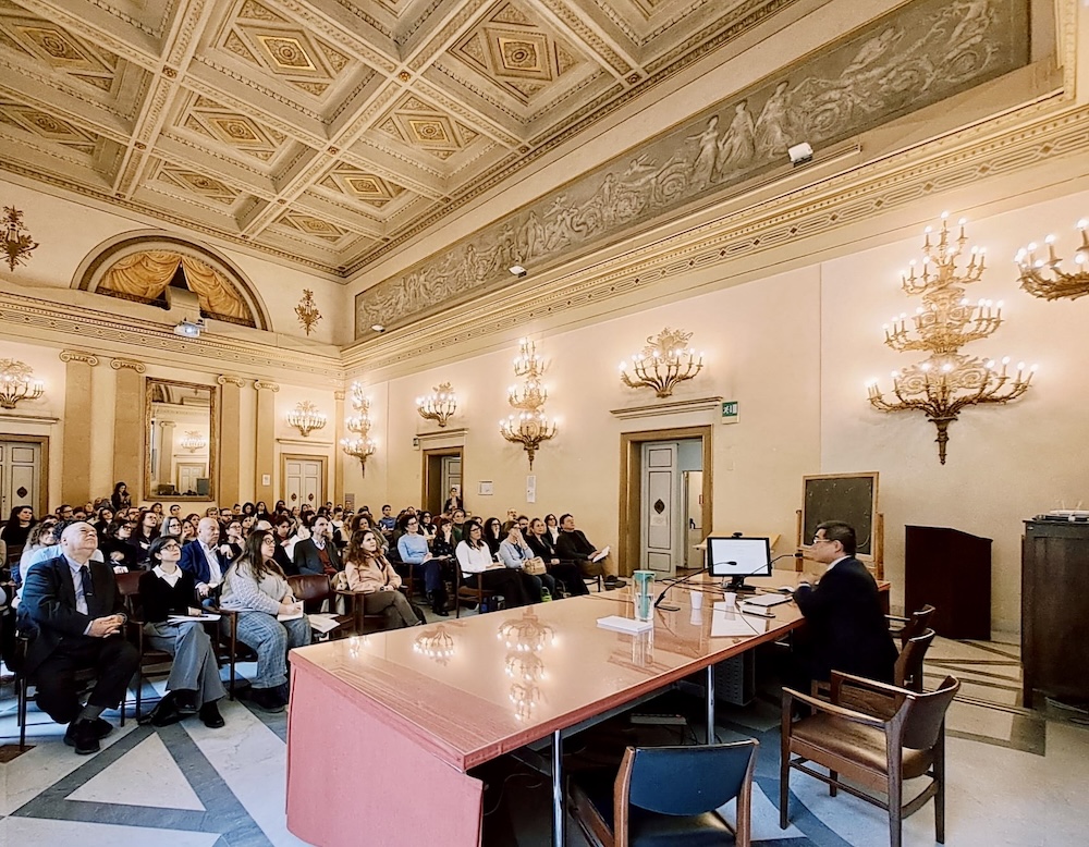 A man sits at a large table, facing an ornate room full of an interested audience. Chandoliers line the room and the ceiling is highly decorated.