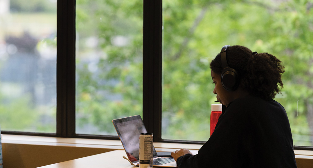 A person with curly hair and headphones works on a laptop at a table next to a large window with a view of greenery. A red water bottle and a white can are on the table.
