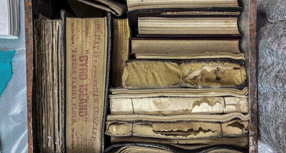 A collection of worn and aged books is stacked vertically and horizontally in a wooden crate. One visible book cover reads "The Haxall-Crenshaw Company" followed by "Byrd Island Patent Family Flour." The books show signs of wear, with some pages and covers exhibiting creases and discoloration.