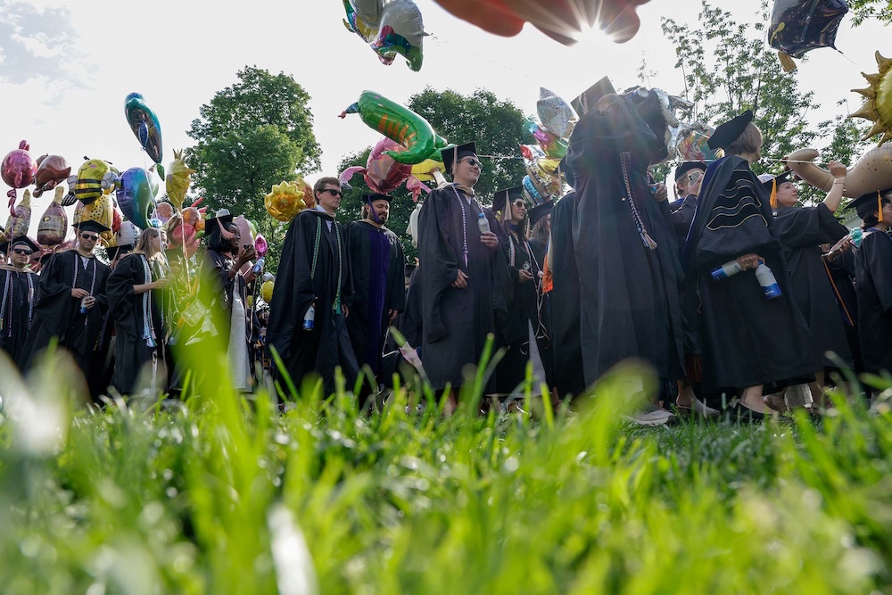 A group of graduates in caps and gowns walk across a grassy area. They are holding colorful balloons, and the sun shines through the trees in the background.