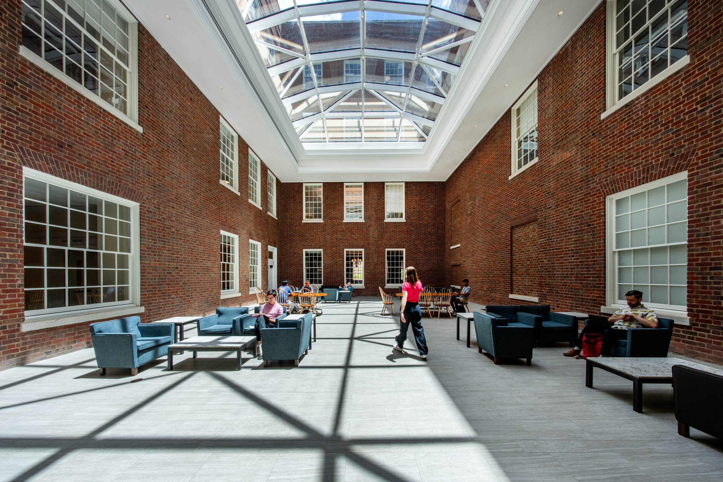 A spacious indoor courtyard with a glass ceiling, brick walls, and large windows. Comfortable seating areas are arranged with chairs and small tables. Natural light floods the area, creating a bright atmosphere. People are casually seated and walking, enjoying the space.