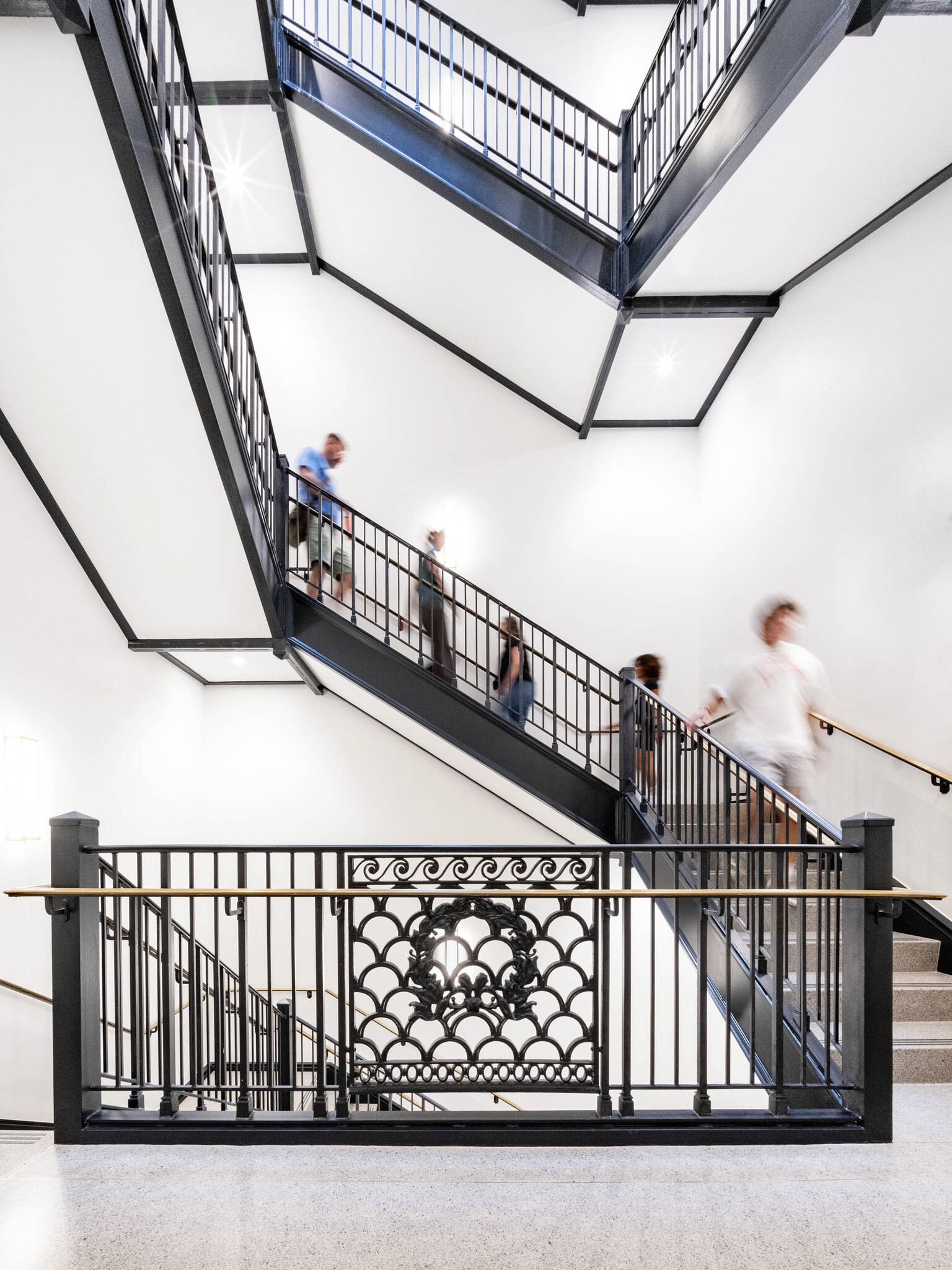 A few people walk up and down a modern, indoor staircase with a decorative wrought iron railing. The stairwell has clean white walls and ample lighting. The scene captures a sense of motion and activity.