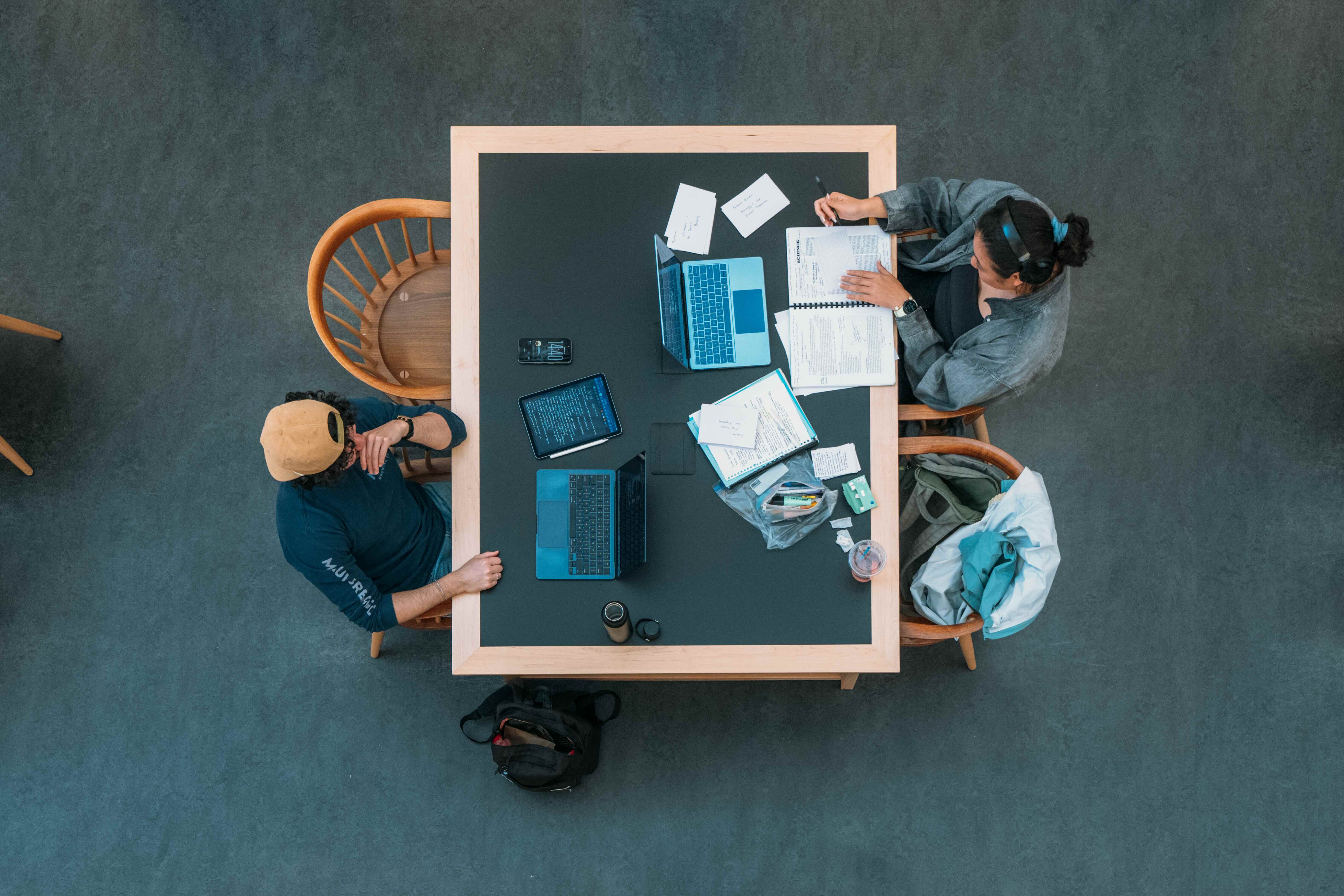 Two people sit across from each other at a table with laptops, papers, and notebooks, viewed from above.