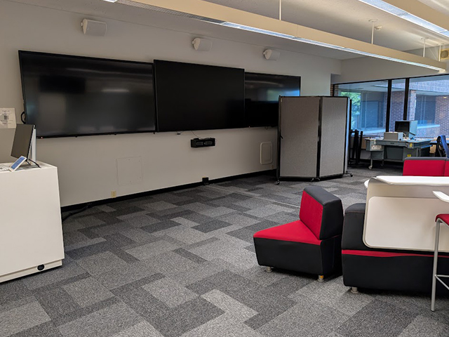 A large horizontal screen with a podium and computer monitor alongside. The large open room also has comfortable red seating.