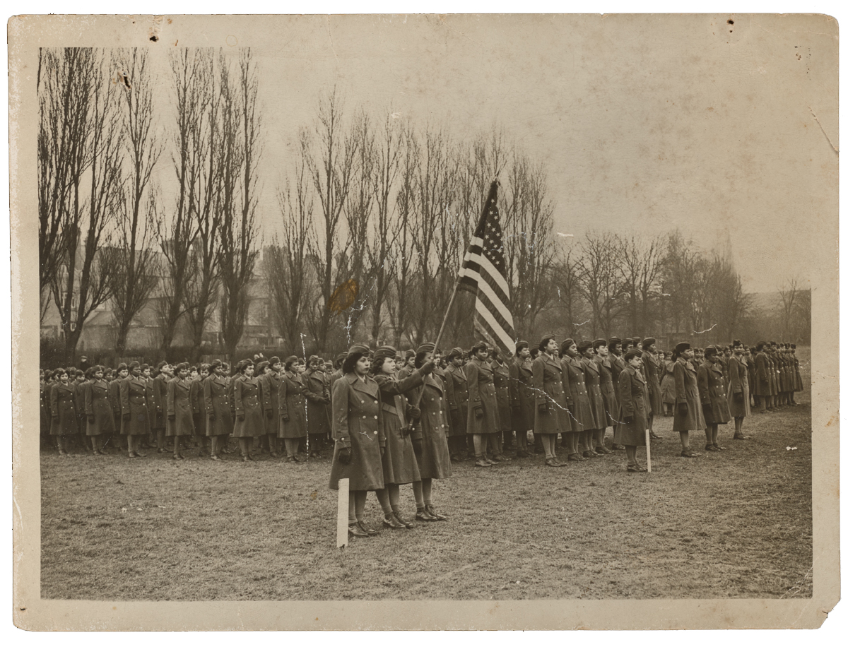 A historic photograph of a military review with female soldiers lined up in formation, holding rifles, and three individuals at the front, one carrying the American flag. 