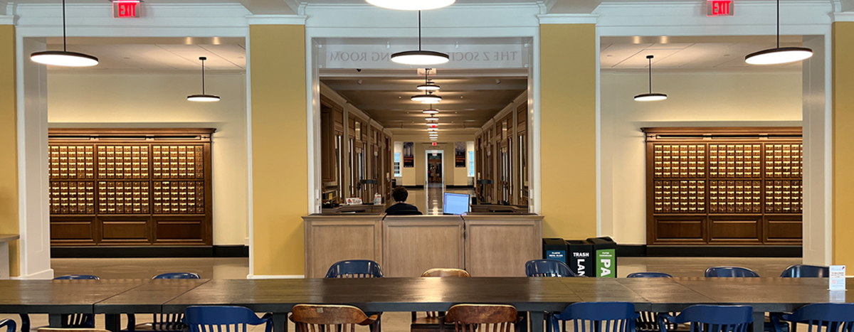 Interior of a library featuring a central service desk with a staff member, rows of card catalog cabinets on other side of a hallway, and multiple tables for study in the foreground.