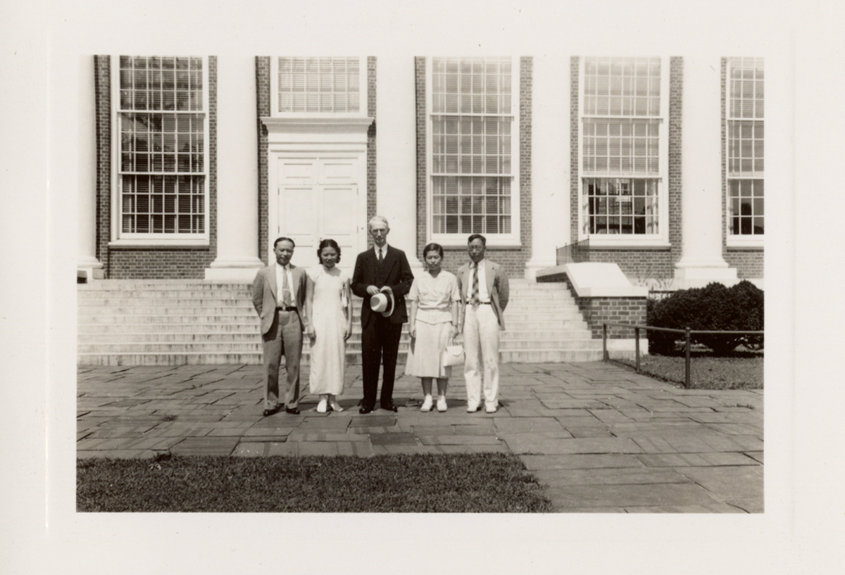 Group of five adults standing in front of the steps of a large brick building with white columns, smiling and dressed in mid-20th century attire.