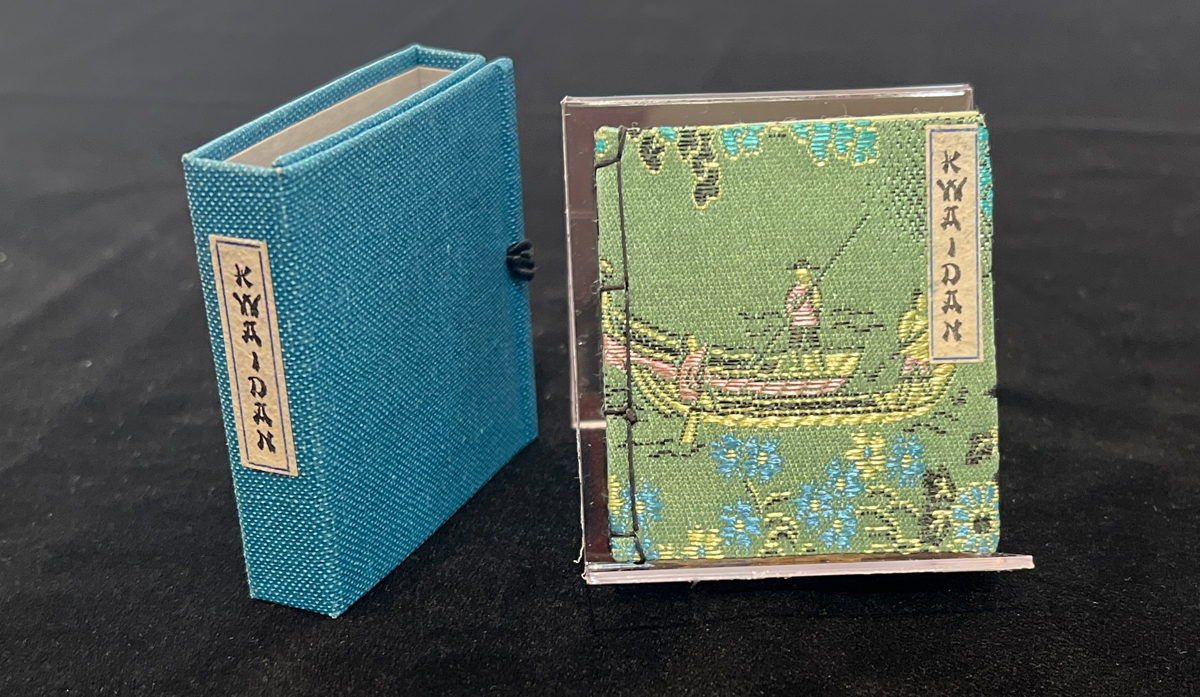 A small, decorative book titled "Kwaidan," displayed with its slipcase with a blue fabric cover side by side on a black surface. The book features a detailed illustration of a traditional Asian scene.