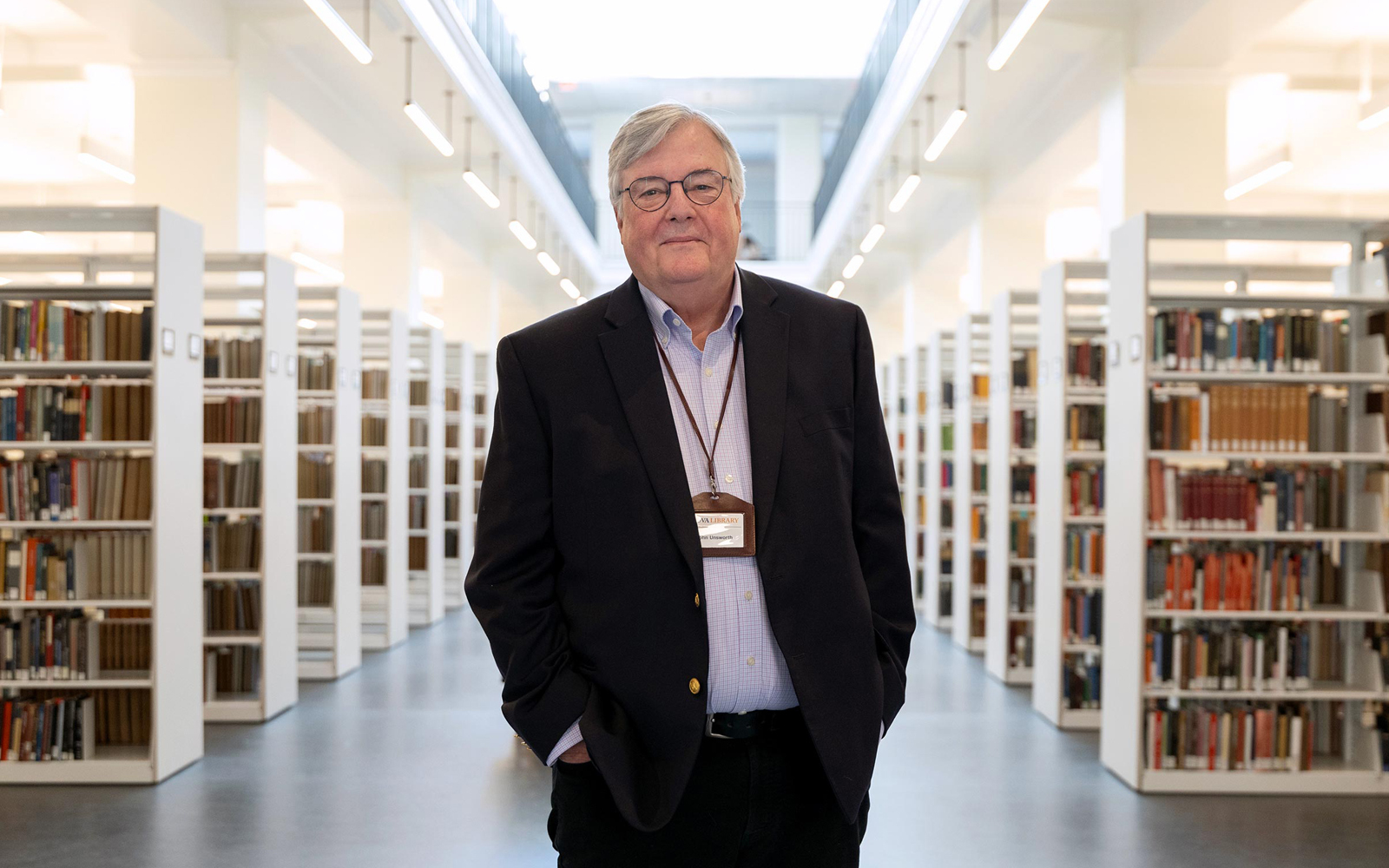 Former UVA Library dean John Unsworth standing in the middle of a library aisle, surrounded by bookshelves stocked with books. He is wearing a dark blazer, light blue shirt, and a badge around his neck.