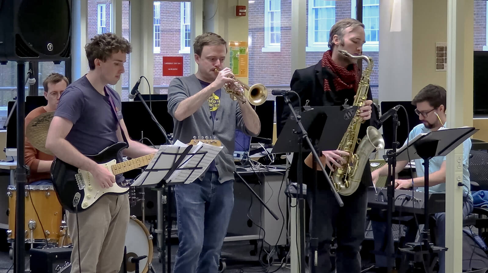 Four musicians perform in a public space, including one on electric guitar, one on trumpet, one on saxophone, and another on keyboard, focused on their sheet music.