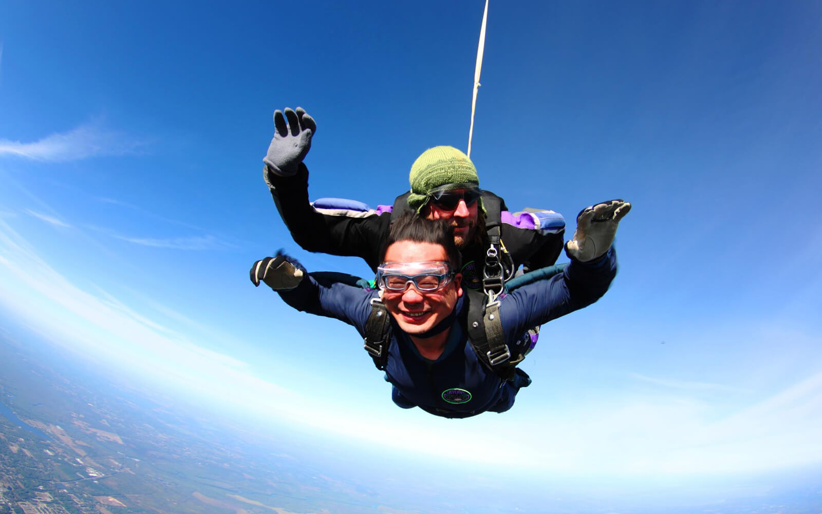 Two people tandem skydiving with a clear blue sky in the background. The person in the front is smiling broadly, and both are wearing protective gear and goggles.