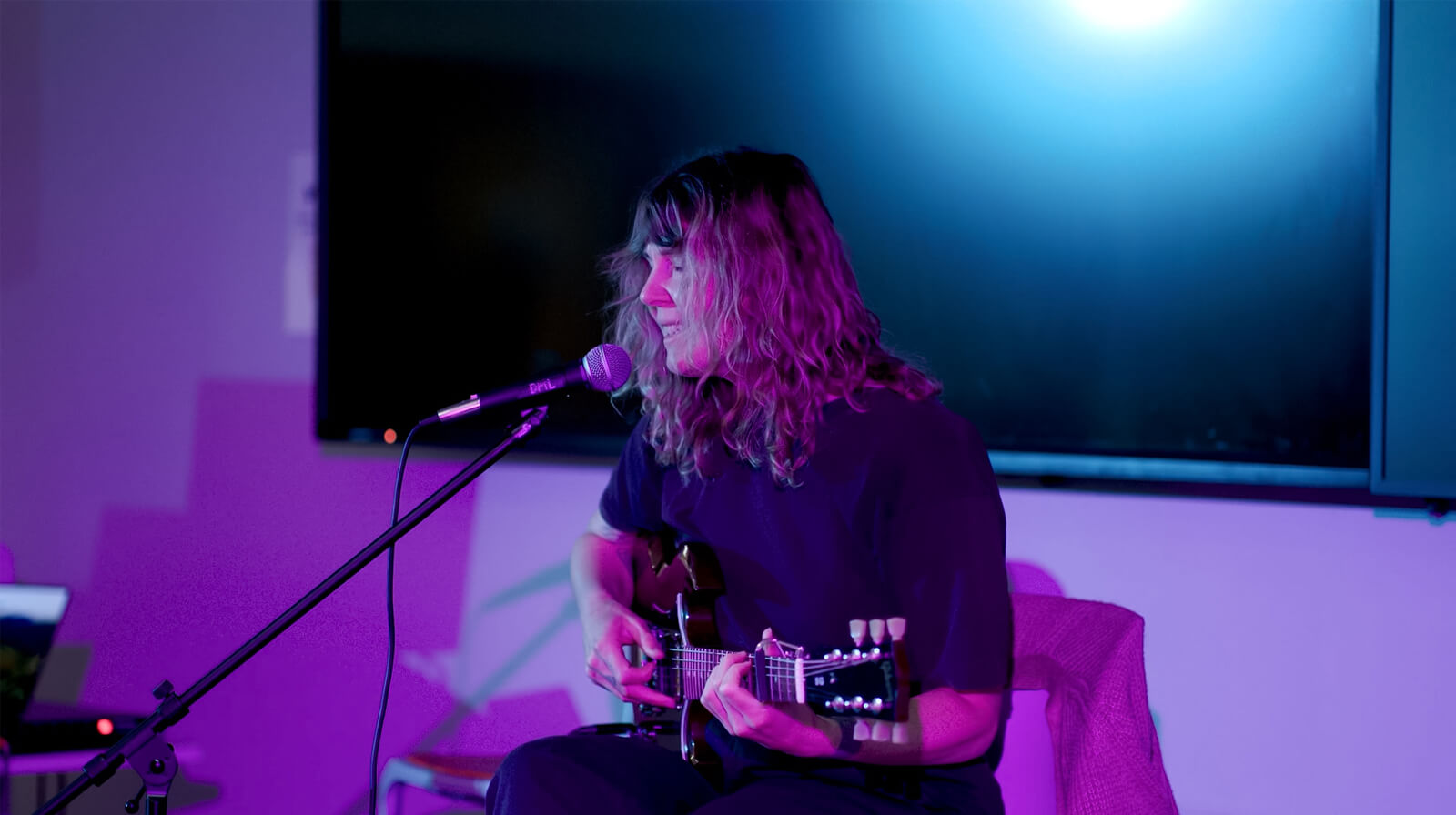Musician performing live in a room with purple lighting, playing an acoustic guitar and singing into a microphone.