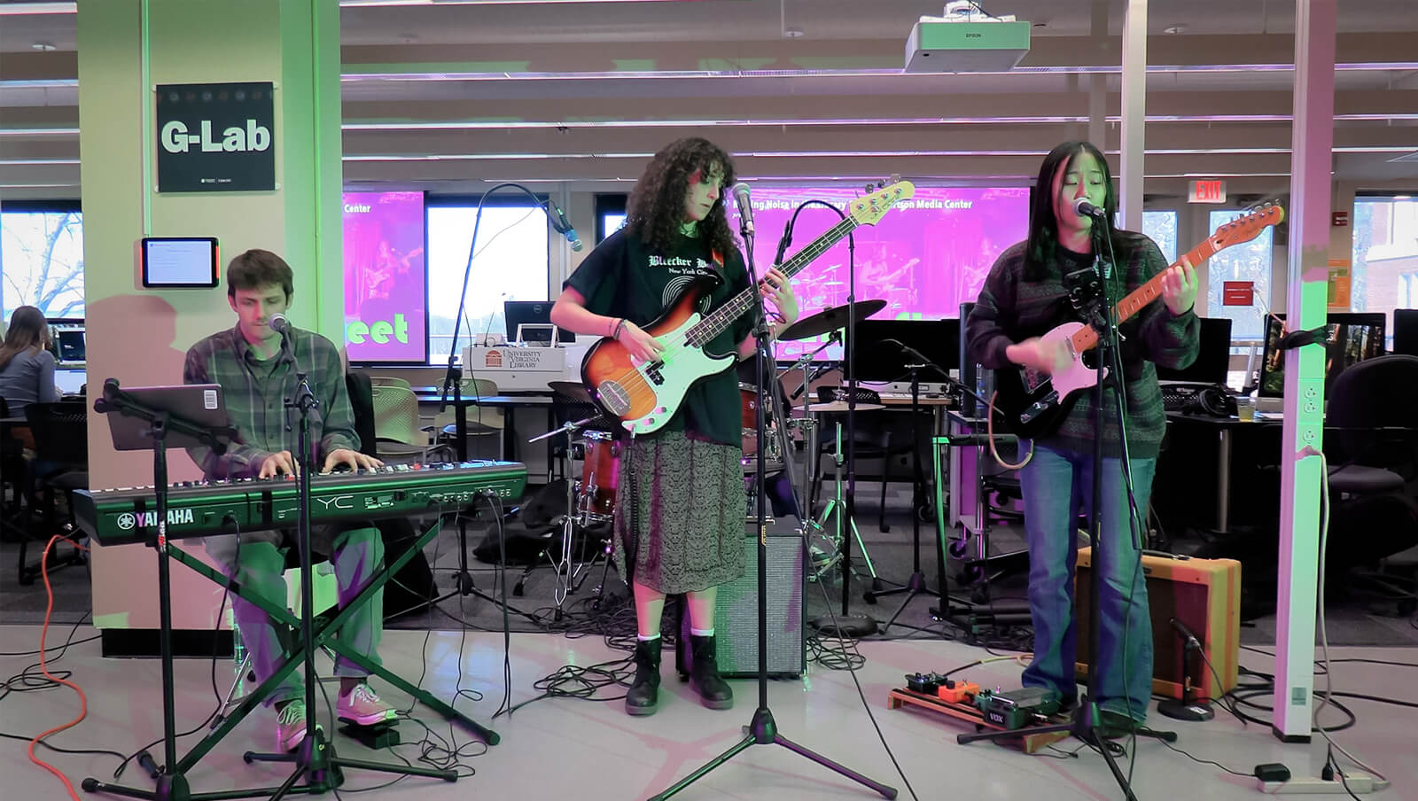 A band performs live inside a room labeled "G-Lab," featuring a keyboard player on the left, a bass guitarist in the center, and a guitarist on the right. A drummer in the background is partially obscured by the bass guitarist.