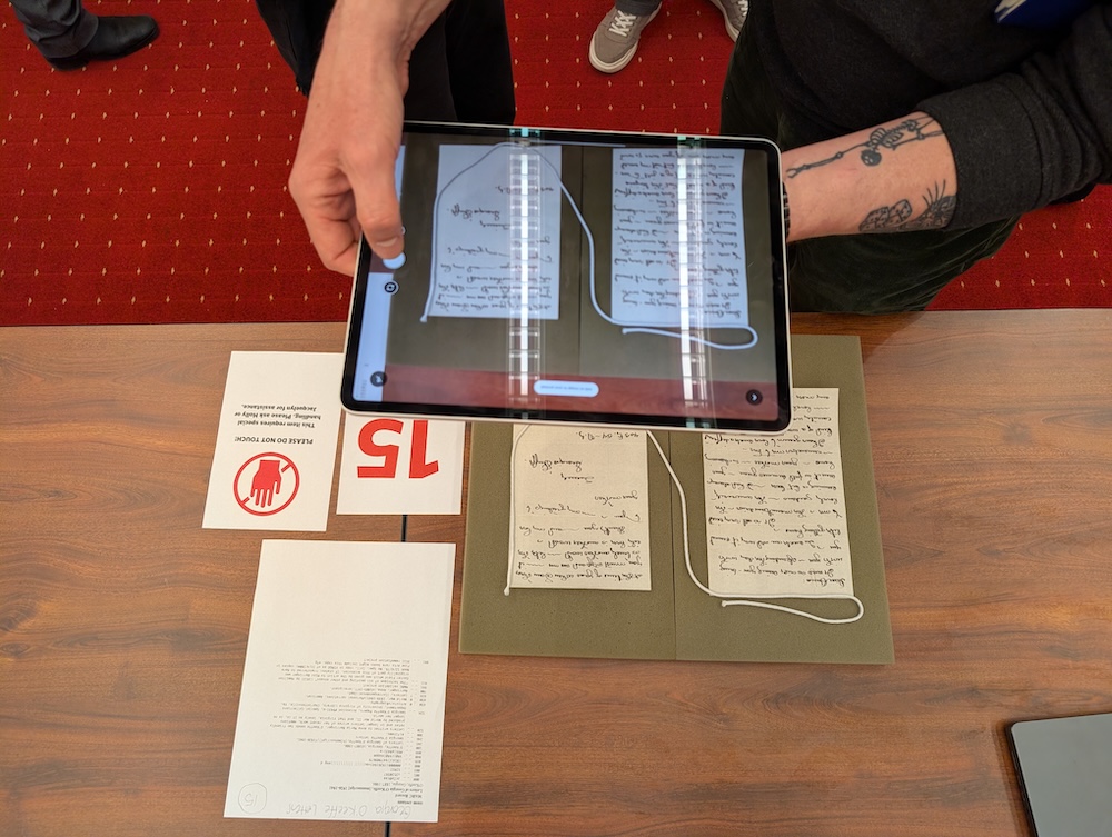 An individual is holding a tablet that scans pages of handwritten text on a table. Nearby, there are printed papers and a sign with a "No touching" symbol.