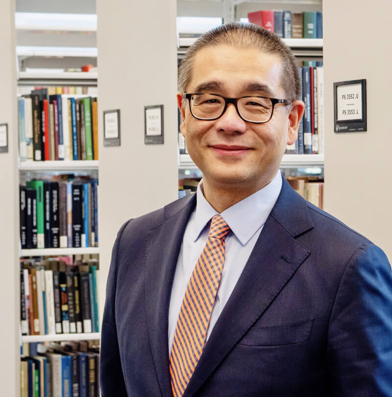 Individual in a business suit and wearing glasses smiling in a library setting, with bookshelves in the background.