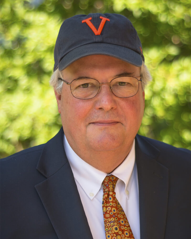 Person wearing a cap with the letter 'V' and a suit, standing outdoors with a background of blurred green foliage.