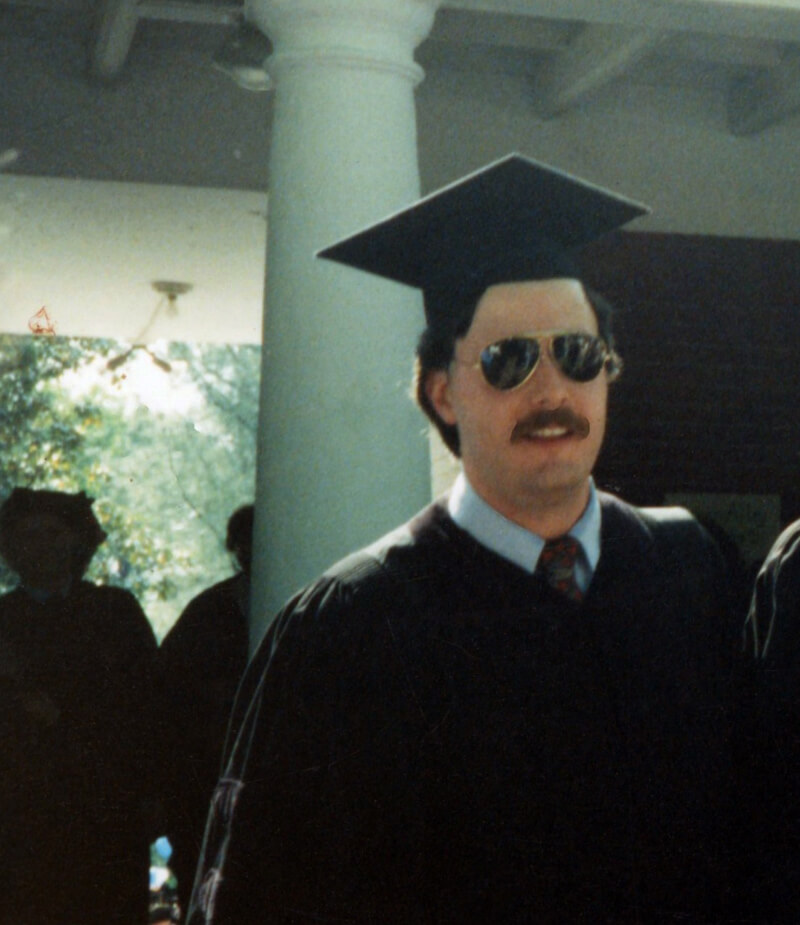 Person in graduation attire with a thick mustache, wearing a cap and sunglasses.