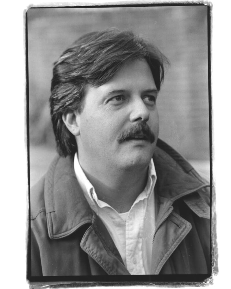 Black and white portrait of a person with a mustache, wearing a jacket over a shirt, looking to the side.