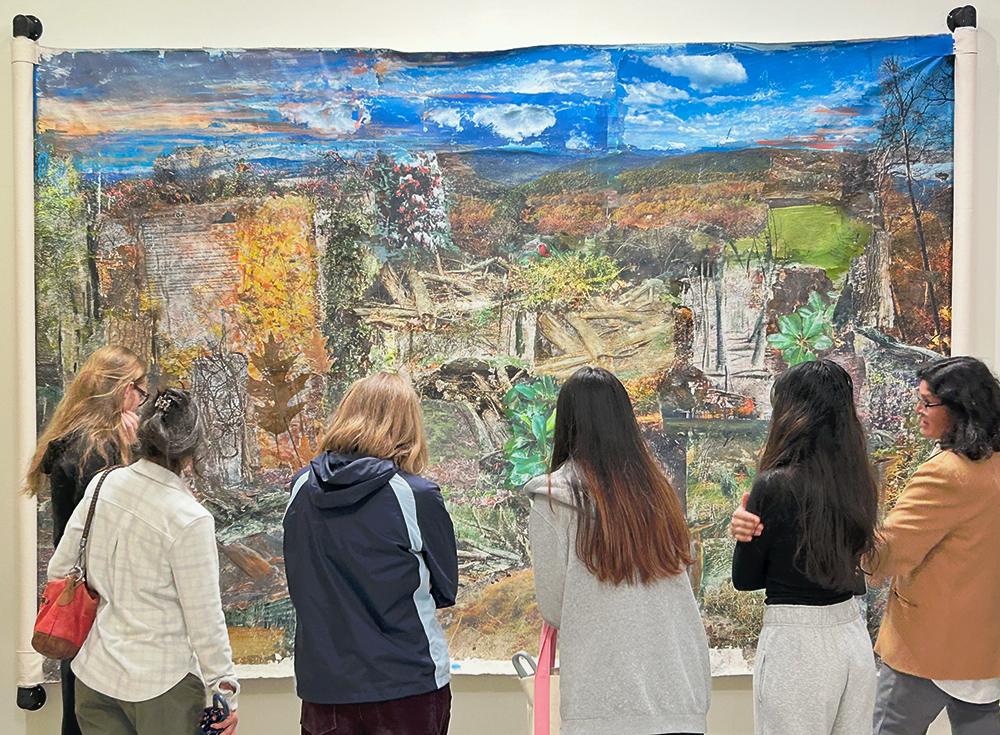 A group of people stand in front of a large scroll, which is stretched across the wall. The canvas scroll is a collage of organic pictures and shapes: leaves, branches, trees, with each shape layering over others.
