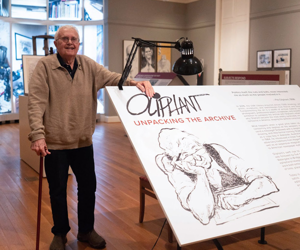 A man with a cane wearing black pants and a tan sweater stands in a exhibition setting. He leans on a large panel which reads "Oliphant: Unpacking the Archive" and shows a drawing of him musing, seating and holding a pen.