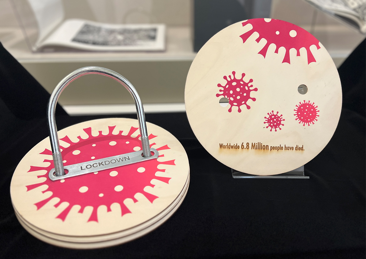 A circular book related to COVID-19 in two pieces. One features a padlock "LOCKDOWN" over a pink virus design. The other is a page detached showing virus illustrations and the text "Worldwide 6.8 Million people have died."