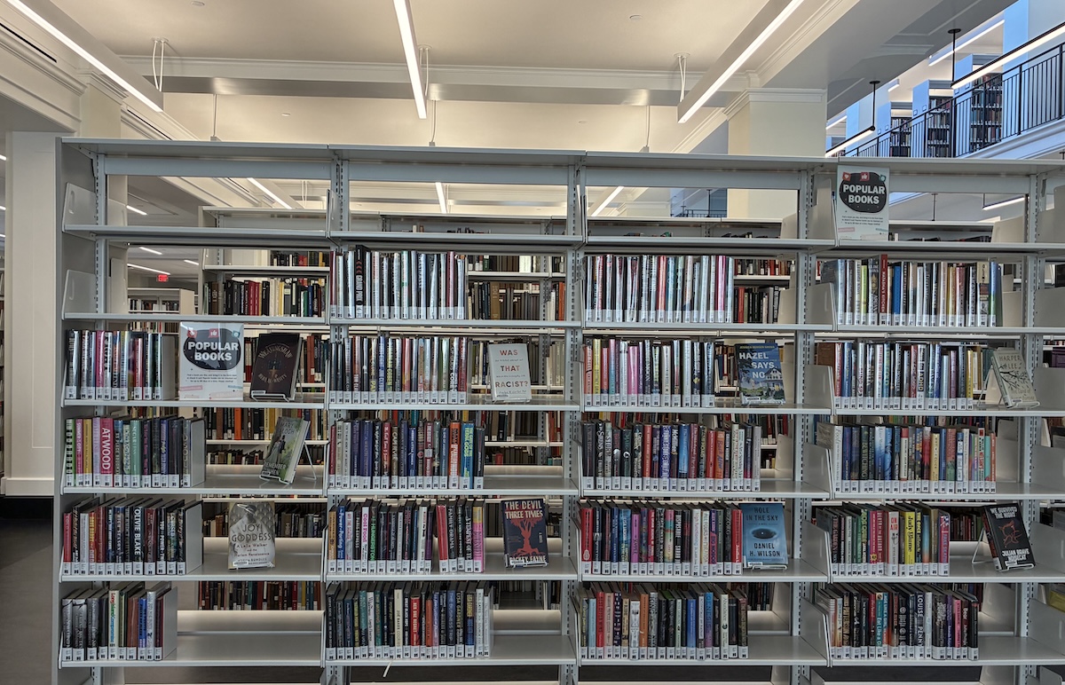 Bookshelves filled with a variety of books are arranged in a library setting. Signs on the shelves say "Popular Books." The room has ample lighting and a modern design, featuring tall windows and metal shelving units.