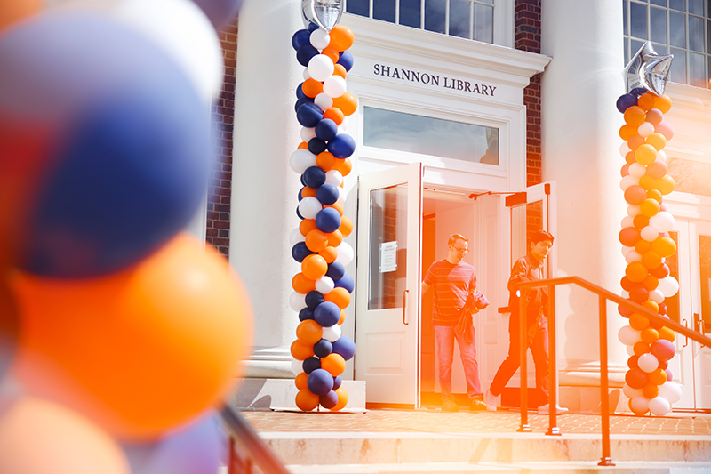 The front door of a building labeled Shannon Library, with smiling people and an orange and blue balloon garland over the door.
