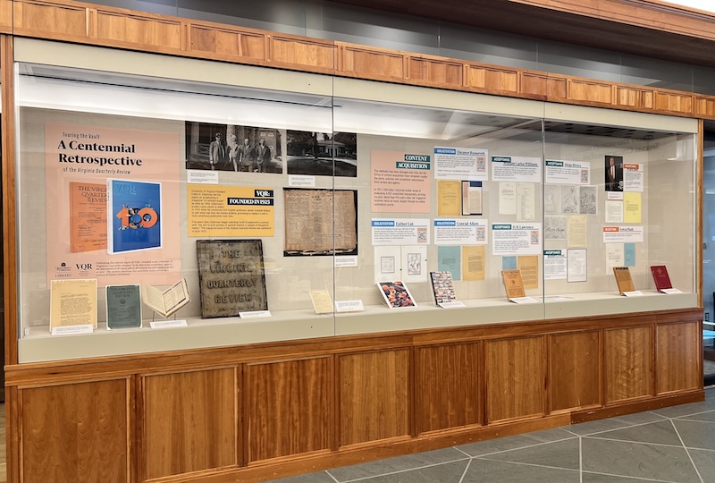 Exhibit display at a library featuring various documents and posters related to a centennial retrospective, housed in a wooden cabinet with glass panes.