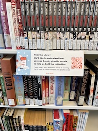 Graphic novels are displayed on library shelves, including series like "Bleach" and volumes with various colorful spines. A sign with a QR code invites patrons to share how they use graphic novels to help build the collection.