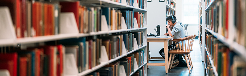 Person wearing headphones studies at a library table between tall bookshelves.
