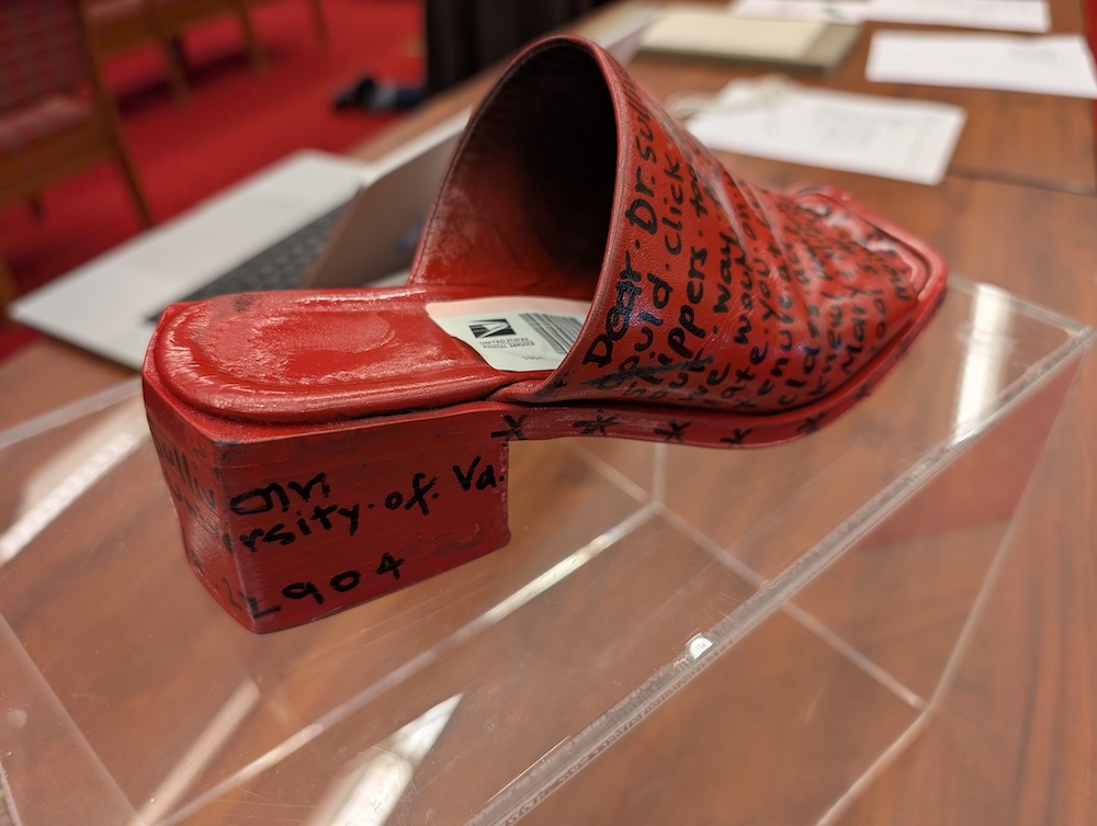 A red shoe displayed on a clear stand. The shoe is covered with handwritten text, including "University of Va." and "1904." In the background, there are papers and a partially visible laptop on a wooden table.