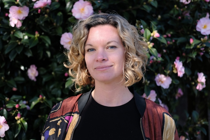 A woman stands, smiling, in front of flowers.